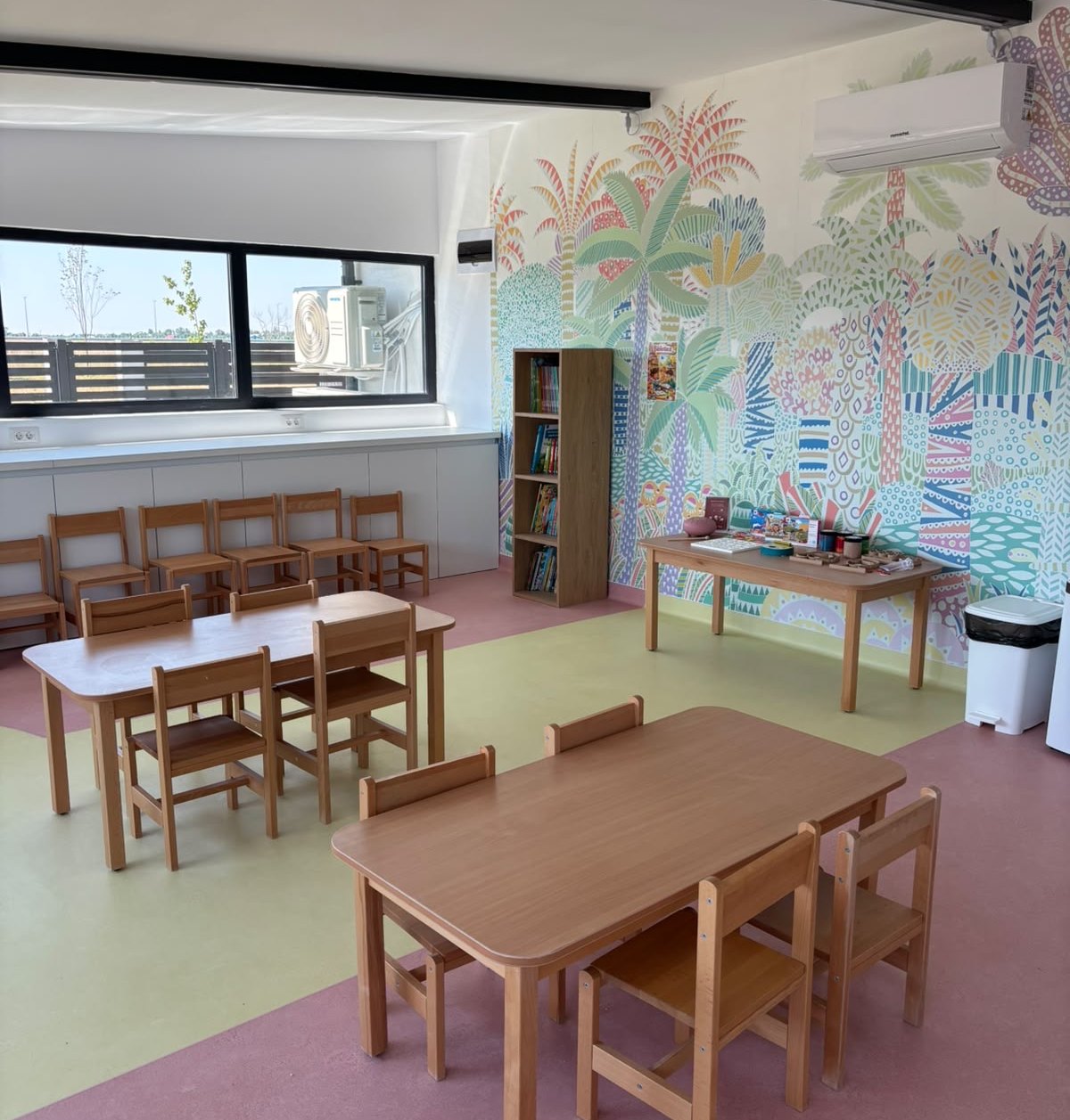 Sala de grupă: mese și scăunele din lemn, bibliotecă și perete pictat